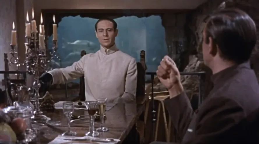 Dr. No hosts Bond for an ominous candlelit dinner in his island lair, aquarium glowing behind him